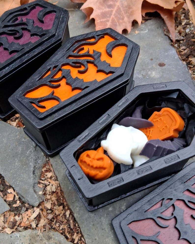 May include: Black coffin-shaped boxes with carved designs and orange, purple, and black interiors. One box is open, revealing Halloween-themed soaps, including a pumpkin, ghost, and bat. The boxes are displayed on a stone surface.