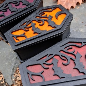May include: Three black coffin-shaped boxes with decorative bat and vine cutouts on the lids. The interiors are colored orange, red, and purple. The boxes are displayed outdoors on a stone surface, with fallen leaves in the background.