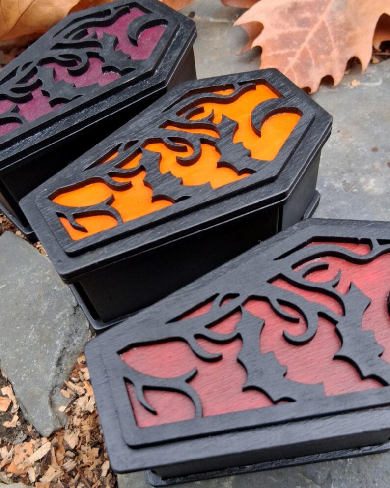 May include: Three black coffin-shaped boxes with decorative bat and vine cutouts on the lids. The interiors are colored orange, red, and purple. The boxes are displayed outdoors on a stone surface, with fallen leaves in the background.