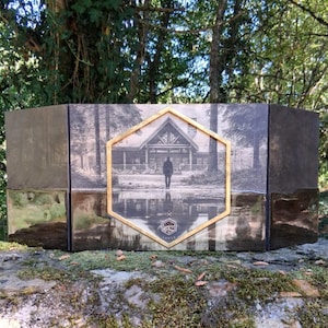May include: A multi-panel decorative box with a gold-framed image of a cabin and a person standing in front of it. The box has a dark, textured exterior with metallic accents. The box is resting on a moss-covered stone surface.