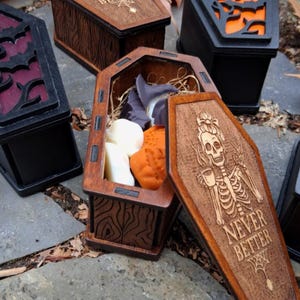 May include: Wooden coffin-shaped boxes with intricate designs, some open to reveal contents. One box features a skeleton with the text "NEVER BETTER." Other boxes have decorative cutouts and are painted black or brown. The open box contains wax melts.