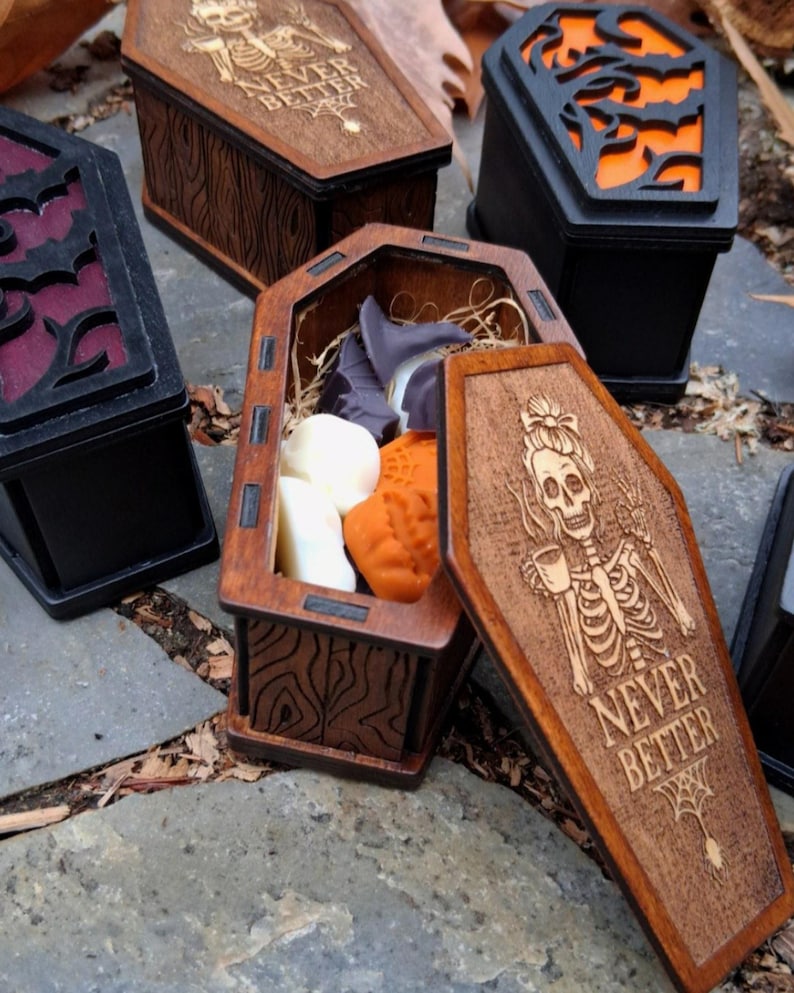 May include: Wooden coffin-shaped boxes with intricate designs, some open to reveal contents. One box features a skeleton with the text "NEVER BETTER." Other boxes have decorative cutouts and are painted black or brown. The open box contains wax melts.