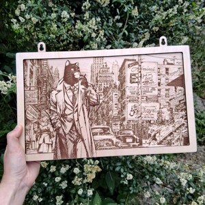 Op de afbeelding: Houten wandkunst met een kat in een pak in een stadsgezicht. Het kunstwerk is een gedetailleerde gravure met een vintage esthetiek, met gebouwen, auto's en figuren. Het stuk heeft twee ophanglussen.