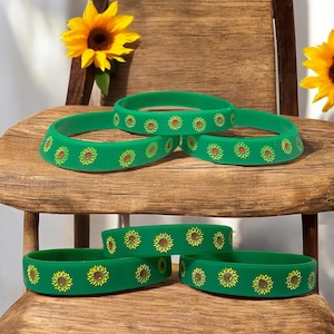 May include: Five green silicone bracelets with a sunflower design. The sunflowers have yellow petals and red centres.