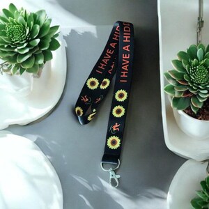 May include: Black lanyard with sunflower and flame designs. The lanyard has the text "I have a hidd..." printed on it.