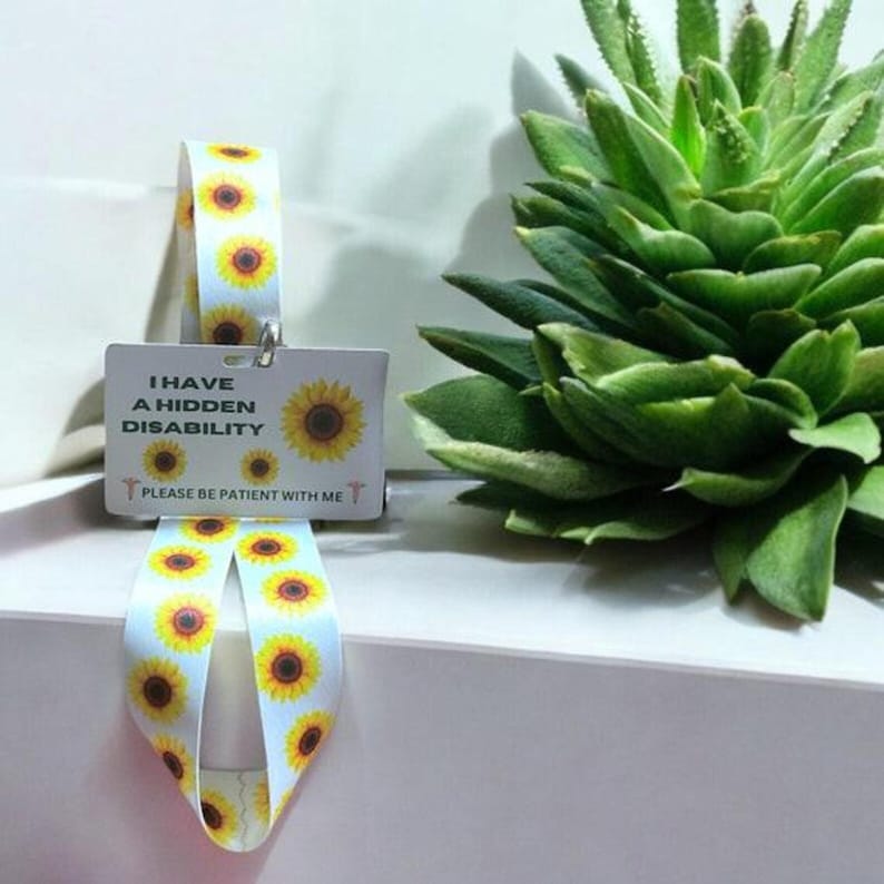 May include: White card with the text "I have a hidden disability. Please be patient with me." and a sunflower graphic. The card is attached to a white ribbon with a sunflower pattern.