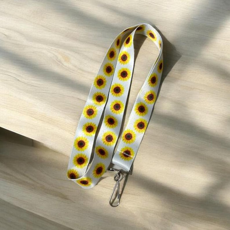 May include: White lanyard with a sunflower pattern. The lanyard has a silver metal clip.