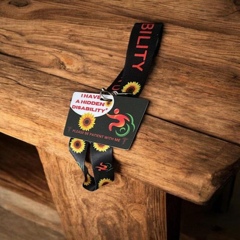 May include: A black lanyard with a sunflower pattern and the word "BILITY" printed in red. Attached to the lanyard is a black card with the text "I HAVE A HIDDEN DISABILITY* PLEASE BE PATIENT WITH ME" and a stylized image of a person in a wheelchair.