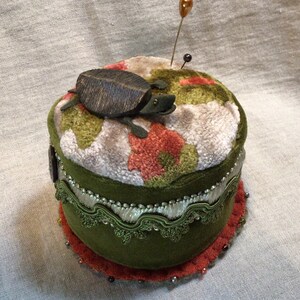 May include: A handcrafted pin cushion with a green velvet base and a floral patterned top. A wooden turtle decorates the top, with two pins inserted. The cushion is trimmed with decorative trim and a beaded edge.