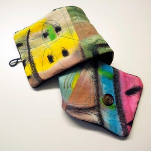 Puede incluir: Dos fundas de tela acolchada con diseños abstractos. Una es amarilla, verde y negra, la otra es azul, rosa y verde. Ambas tienen ribetes negros y costuras azules. Una tiene cierre de botón y la otra un lazo.