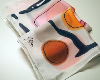 May include: Three folded silk scarves with abstract designs. The scarves feature orange, pink, and navy blue shapes on a white background. One scarf has the signature "Gilben" in black. The scarves are neatly folded and stacked.