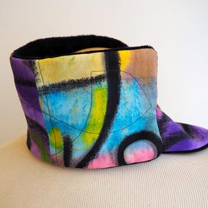 May include: A brightly coloured neck warmer with an abstract design. The fabric displays a mix of purple, blue, yellow, and pink, with black lines. The interior is lined with black material.