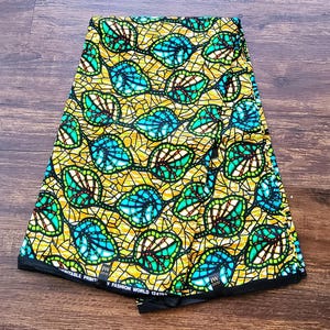 Puede incluir: Un trozo de tela amarilla doblado con un patrón de hojas negras, verdes y azules. La tela tiene el texto "FW FASHION WORLD 1247" impreso en ella.