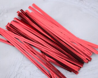 May include: A collection of shiny, red metallic twist ties. These thin, rectangular strips are arranged in a loose pile, reflecting light. They are likely used for securing gift wrapping or crafting projects.