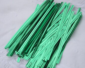 May include: A pile of shiny, metallic green twist ties. These thin, rectangular strips are arranged in a diagonal pattern, reflecting light. The ties are likely used for gift wrapping or crafting.