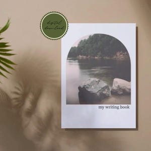 Peut inclure: Un carnet d'écriture rectangulaire à couverture blanche orné d'un graphique en forme d'arche. L'image à l'intérieur de l'arche montre une rivière calme, de gros rochers au premier plan et une forêt dense. Le texte my writing book est imprimé.