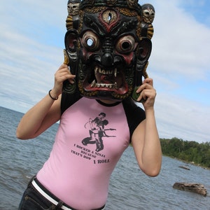 May include: A black and red mask with multiple faces on top is held up in front of a person. The person is wearing a pink and black shirt with the text "I rocked a ninja because that's how I roll". The person is wearing blue jeans and a white belt.