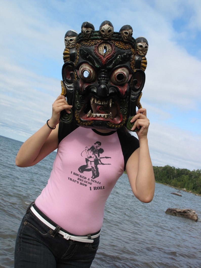 May include: A black and red mask with multiple faces on top is held up in front of a person. The person is wearing a pink and black shirt with the text "I rocked a ninja because that's how I roll". The person is wearing blue jeans and a white belt.