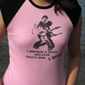 May include: Pink and black raglan t-shirt with a graphic of a guitarist and the text "I rocked a ninja because that's how I roll". The shirt is made of a soft material.