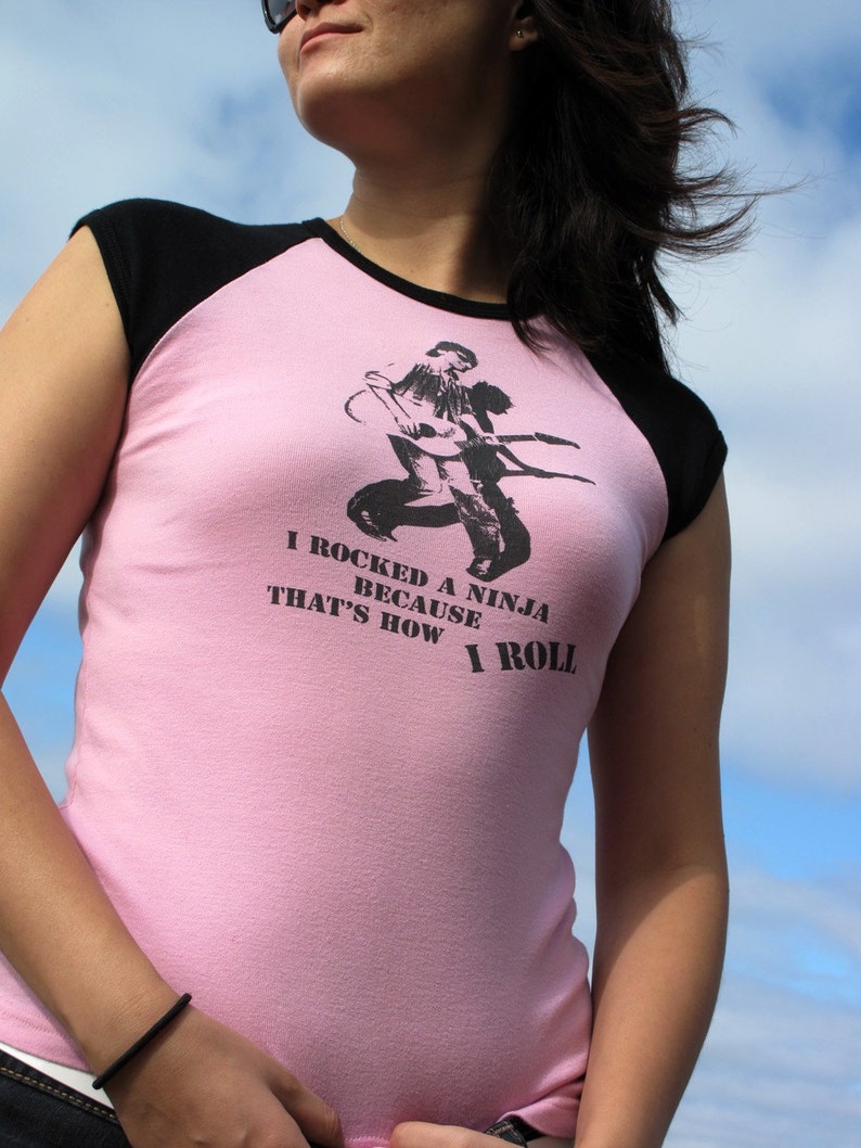 May include: Pink and black raglan t-shirt with the text "I rocked a ninja because that's how I roll" and a black graphic of a ninja playing a guitar. The shirt has black sleeves and a pink body.