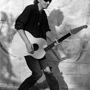 May include: Black and white image of a person playing an acoustic guitar. The guitar is light-colored with a dark fretboard. The person is wearing sunglasses, a black shirt, and ripped jeans. The background is a brick wall.