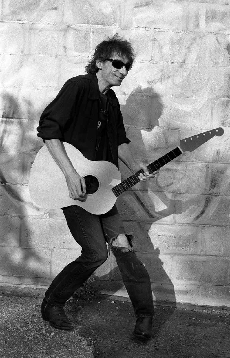 May include: Black and white image of a person playing an acoustic guitar. The guitar is light-colored with a dark fretboard. The person is wearing sunglasses, a black shirt, and ripped jeans. The background is a brick wall.