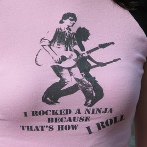 May include: Pink raglan t-shirt with black sleeves and collar. The shirt features a black and white graphic of a person playing a guitar, with the text "I rocked a ninja because that's how I roll."
