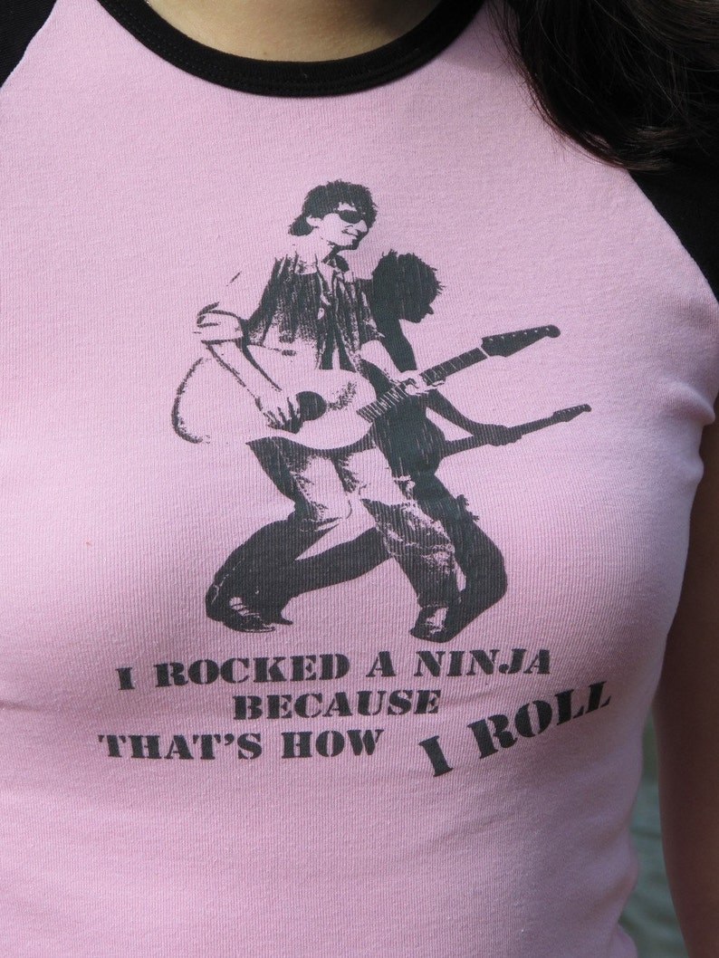 May include: Pink raglan t-shirt with black sleeves and collar. The shirt features a black and white graphic of a person playing a guitar, with the text "I rocked a ninja because that's how I roll."