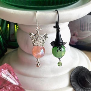 May include: A pair of whimsical earrings. One earring features a silver crown with a pink orb and star charm. The other earring has a black witch hat atop a green orb with a leaf design and a green bead charm. The word "WICKED" is visible in the background.