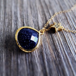 Pode incluir: Um colar dourado com um pingente de pedra preciosa azul escuro facetada. O pingente tem formato de gota e &eacute; cravejado em uma moldura dourada. Um pequeno pingente de estrela dourada com a letra "M" pende perto do fecho.