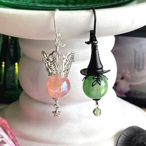 May include: Two unique earrings. One features a silver crown above a pink orb with a star charm. The other has a black witch hat atop a green orb with a leaf design and a green charm. The earrings are displayed on a white pedestal.