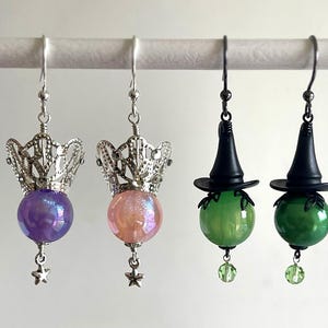 May include: Earrings featuring a variety of designs. Two pairs have silver filigree crowns above iridescent purple and pink orbs, with star charms. The other two pairs have black witch hats above green orbs, with green bead charms.