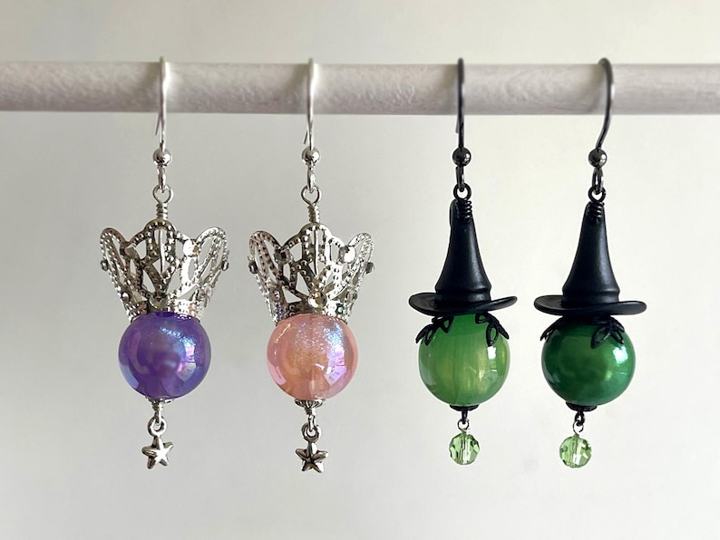 May include: Earrings featuring a variety of designs. Two pairs have silver filigree crowns above iridescent purple and pink orbs, with star charms. The other two pairs have black witch hats above green orbs, with green bead charms.