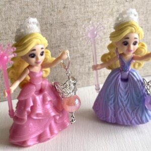 May include: Two princess figurines with blonde hair and tiaras, wearing pink and purple gowns. Each holds a wand and a dangling earring with a crown charm and a gemstone. The earrings have a pink and purple gemstone.