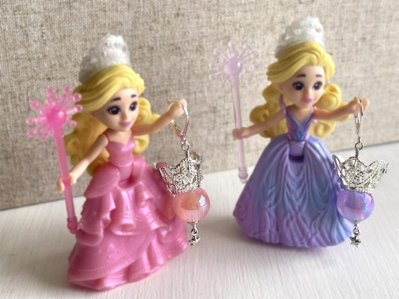 May include: Two princess figurines with blonde hair and tiaras, wearing pink and purple gowns. Each holds a wand and a dangling earring with a crown charm and a gemstone. The earrings have a pink and purple gemstone.