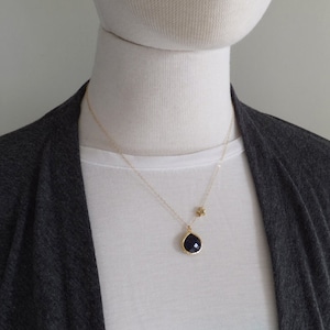 Pode incluir: Um colar de ouro com um pingente em forma de gota azul escuro e um pequeno berloque de estrela. O pingente &eacute; emoldurado em ouro. O colar &eacute; usado sobre uma camiseta branca e um cardigan cinza.