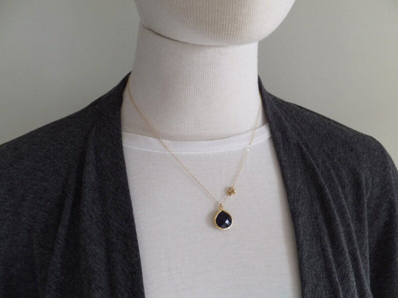 Pode incluir: Um colar de ouro com um pingente em forma de gota azul escuro e um pequeno berloque de estrela. O pingente &eacute; emoldurado em ouro. O colar &eacute; usado sobre uma camiseta branca e um cardigan cinza.