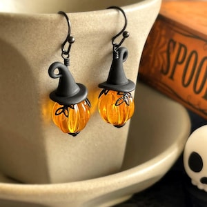 May include: A pair of dangle earrings featuring translucent orange glass pumpkins topped with matte black witch hats. The earrings are suspended from dark metal hooks and include small black wire details resembling bows at the pumpkin stems.