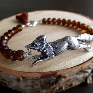 Könnte beinhalten: Ein silbernes Armband mit einem Fuchs-Anhänger und braunen facettierten Glasperlen. Der Fuchs ist in einer laufenden Pose mit offenem Maul dargestellt.