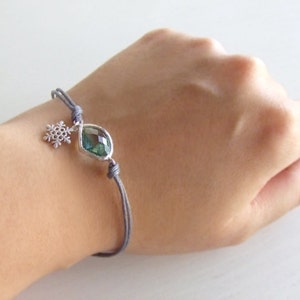 May include: A delicate bracelet featuring a faceted, iridescent gemstone and a snowflake charm. The bracelet is strung on a gray cord and is secured with adjustable knots. The gemstone is approximately 1 cm in length.