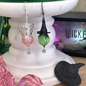 May include: A pair of whimsical earrings. One features a silver crown with a pink iridescent bead and a star charm. The other has a black witch hat over a green bead. A small black witch hat sits below.