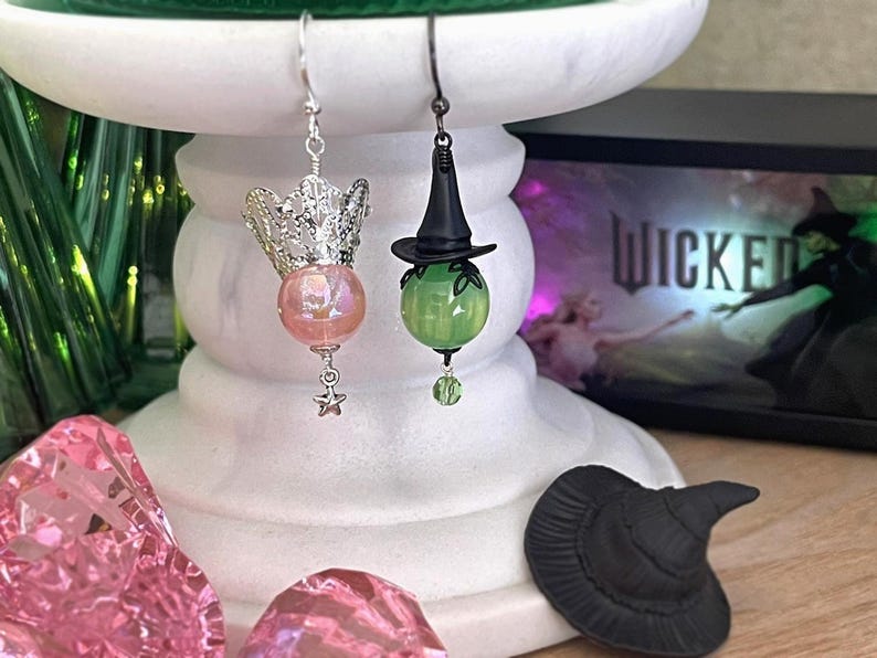 May include: A pair of whimsical earrings. One features a silver crown with a pink iridescent bead and a star charm. The other has a black witch hat over a green bead. A small black witch hat sits below.