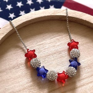 Op de afbeelding: Een zilverkleurige ketting met afwisselend rode stervormige kralen, blauwe stervormige kralen en ronde zilverkleurige kralen bedekt met heldere kristallen. De ketting is in een boogvorm gerangschikt met een patriottisch kleurenschema.