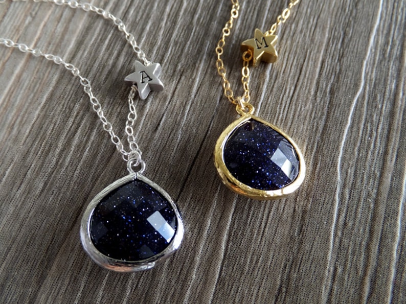 Pode incluir: Dois colares com pingentes em forma de gota. Um tem corrente e moldura prateadas, o outro corrente e moldura douradas. Ambos os pingentes s&atilde;o azul escuro com inclus&otilde;es brilhantes. Cada colar tem um pequeno pingente de estrela com uma letra.