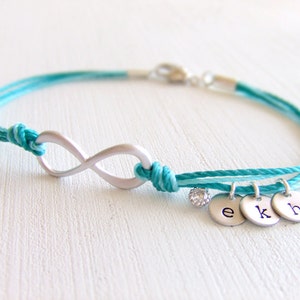 May include: A turquoise and silver infinity bracelet. The bracelet features a silver infinity symbol, a small crystal, and three small round charms with the letters "e", "k", and "h". The bracelet is made of turquoise cord.