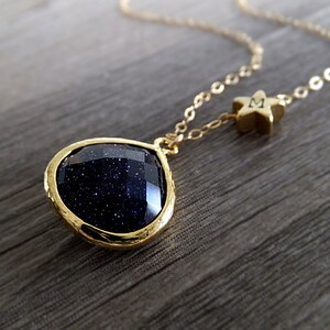 Pode incluir: Colar dourado com um pingente de pedra preciosa azul escuro em uma moldura dourada. H&aacute; tamb&eacute;m um pequeno pingente de estrela dourada com a letra "M". O colar &eacute; exibido em uma superf&iacute;cie de madeira.
