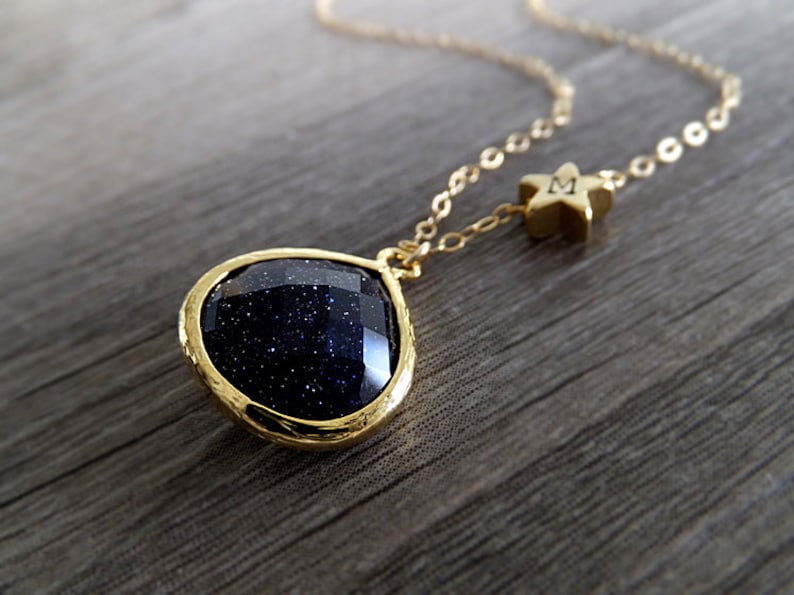 Pode incluir: Colar dourado com um pingente de pedra preciosa azul escuro em uma moldura dourada. H&aacute; tamb&eacute;m um pequeno pingente de estrela dourada com a letra "M". O colar &eacute; exibido em uma superf&iacute;cie de madeira.