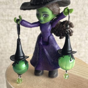 May include: A figurine of a green-skinned witch in a purple dress and black hat, holding two earrings. Each earring features a green orb with a miniature black witch hat and a green crystal charm. The witch is standing on a wooden surface.