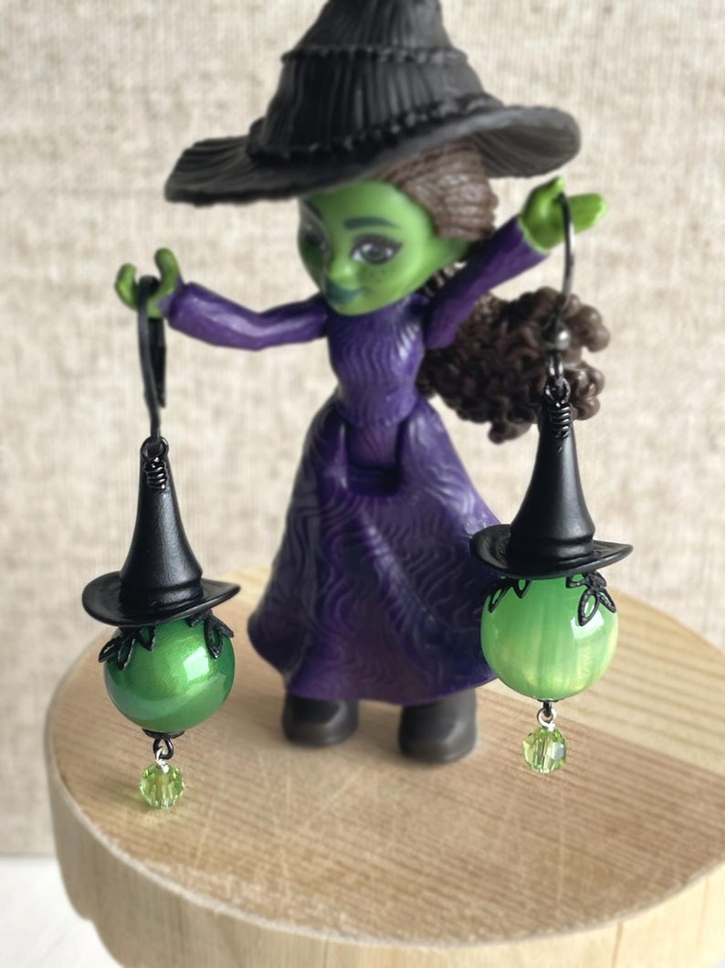 May include: A figurine of a green-skinned witch in a purple dress and black hat, holding two earrings. Each earring features a green orb with a miniature black witch hat and a green crystal charm. The witch is standing on a wooden surface.