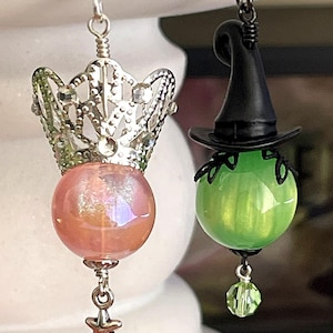 May include: A pair of whimsical earrings. One features a silver crown over a pink iridescent orb with a star charm. The other has a black witch hat atop a green orb with a green crystal charm. Both have silver hooks.